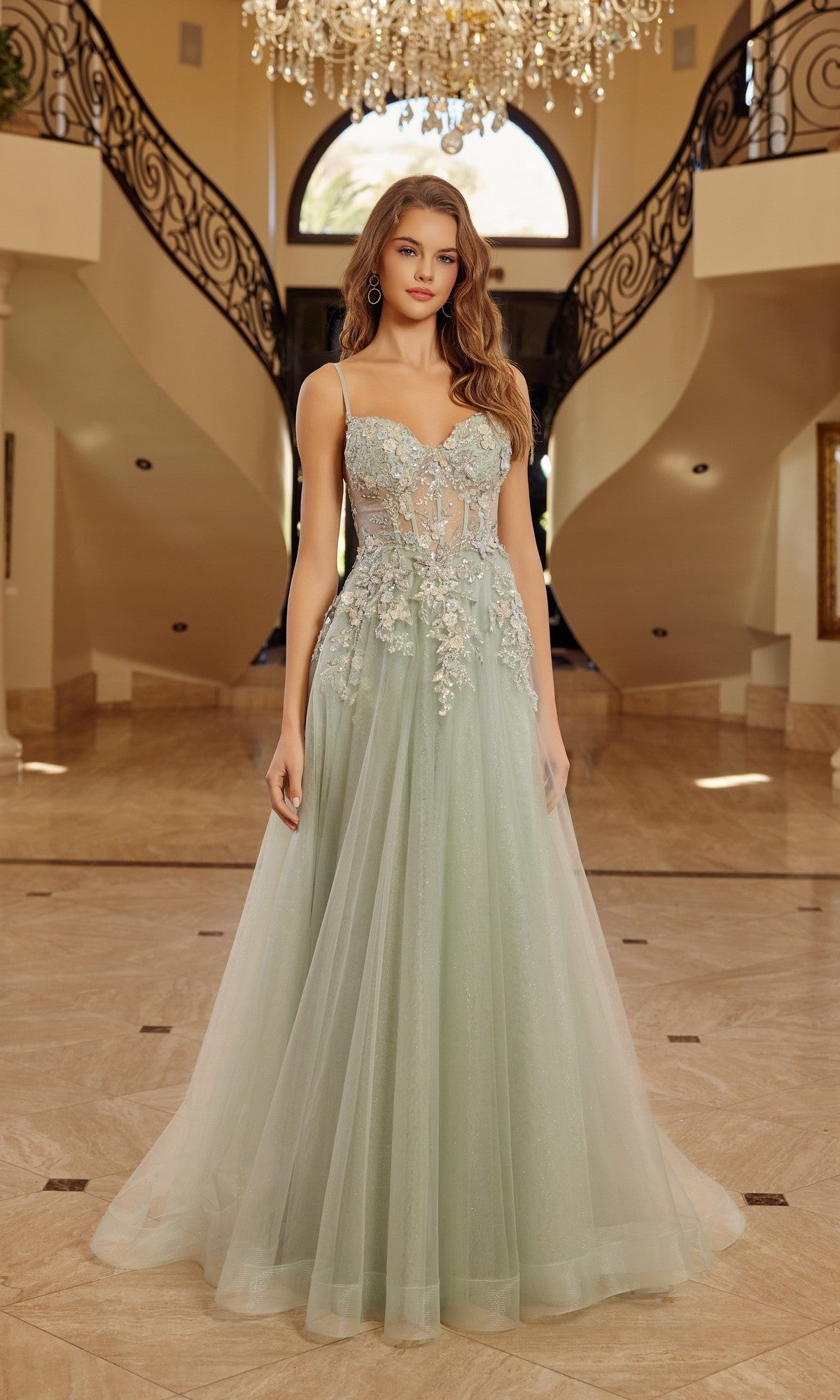 A woman stands in a grand foyer under a chandelier, wearing the Nox Anabel G1787 long prom dress—light green, sleeveless, with sheer details, an elegant A-line silhouette, and delicate 3D floral appliques.